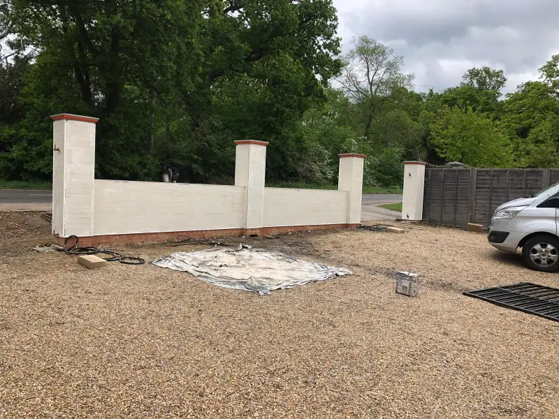 Long rendered boundary wall with three pillars beside a gravel driveway